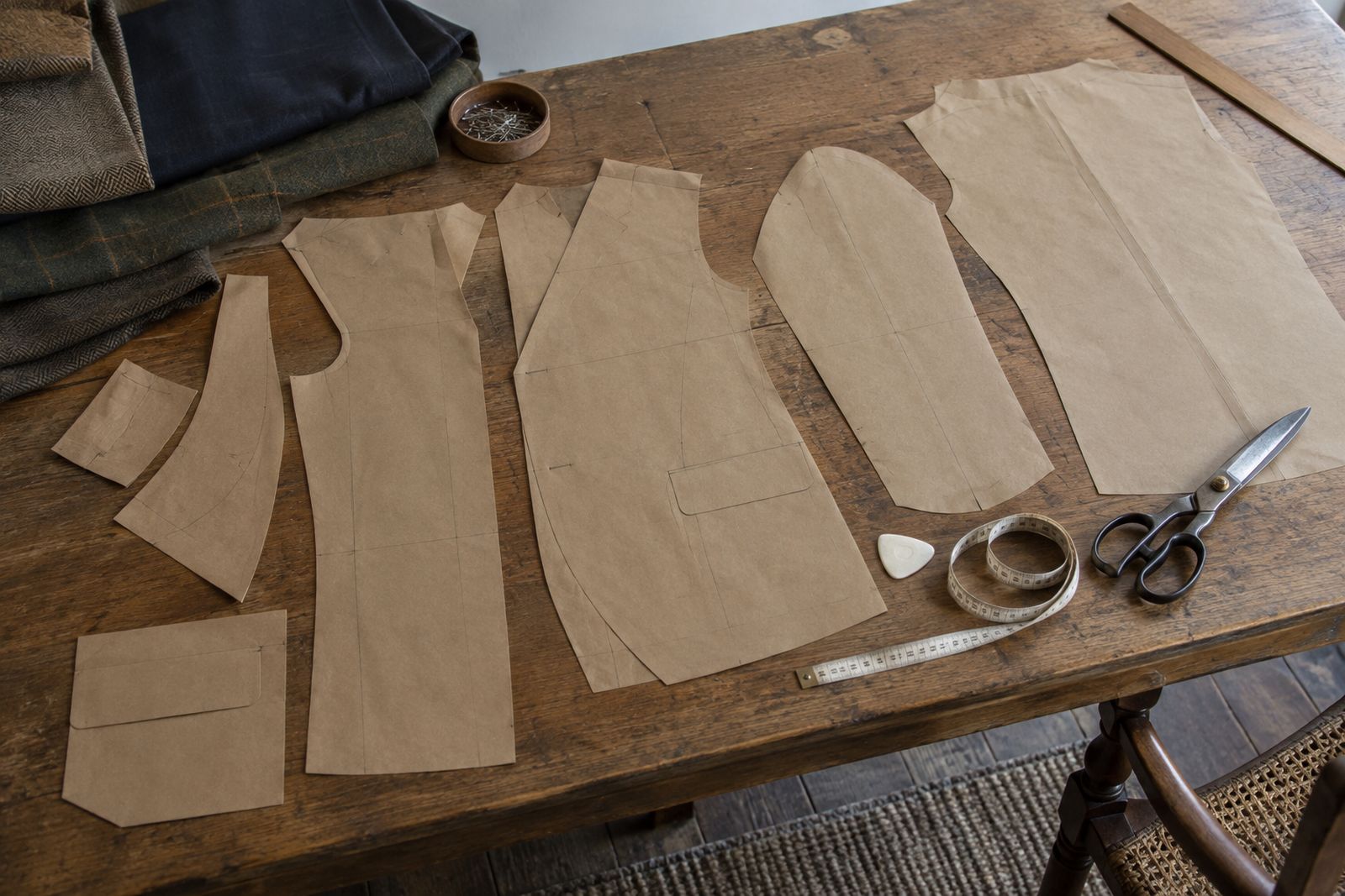 Brown paper jacket pattern pieces on a cutting table beside tailor's chalk, a tape measure and shears.