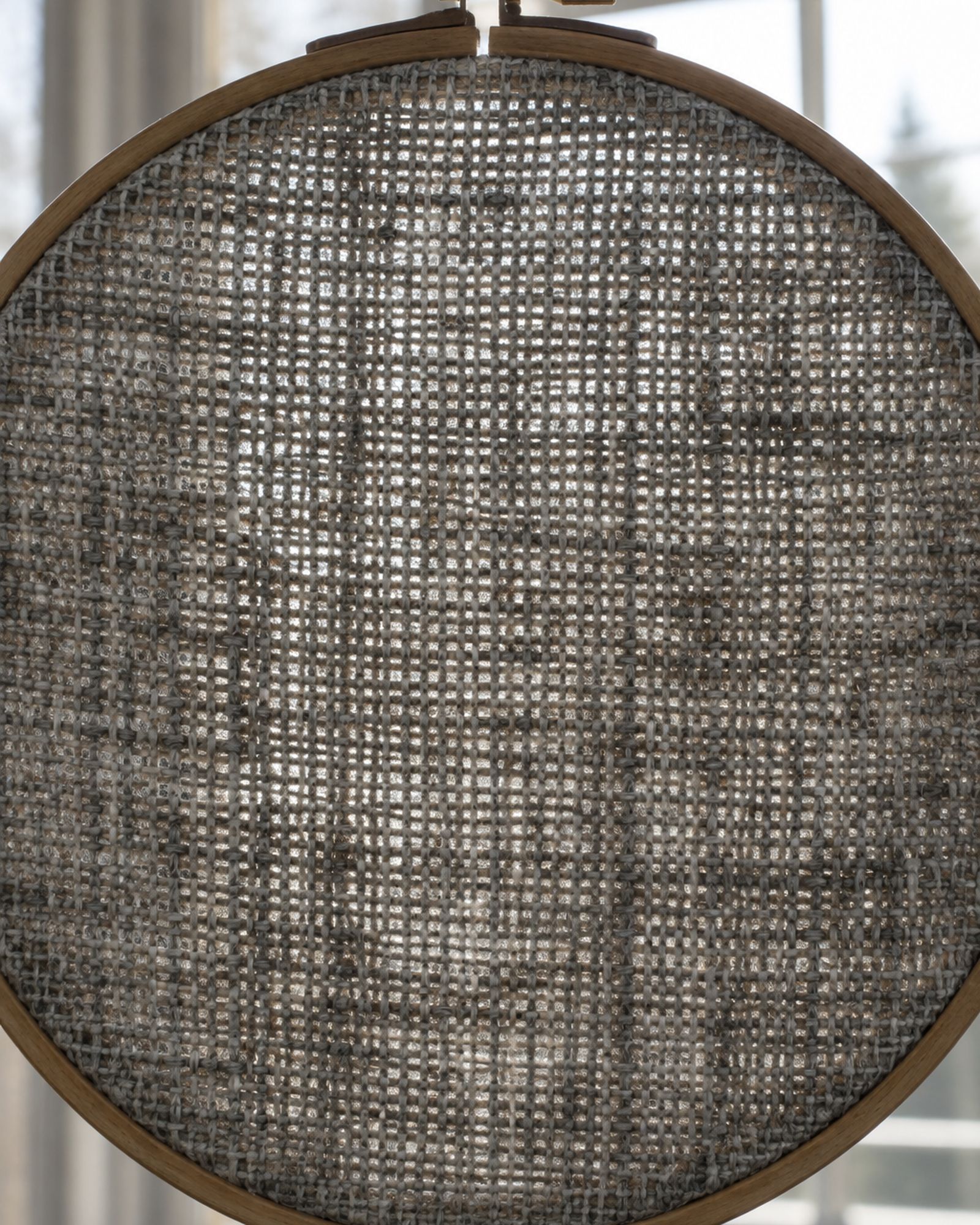 Daylight showing through the open weave of a gray wool swatch held in a wooden hoop.