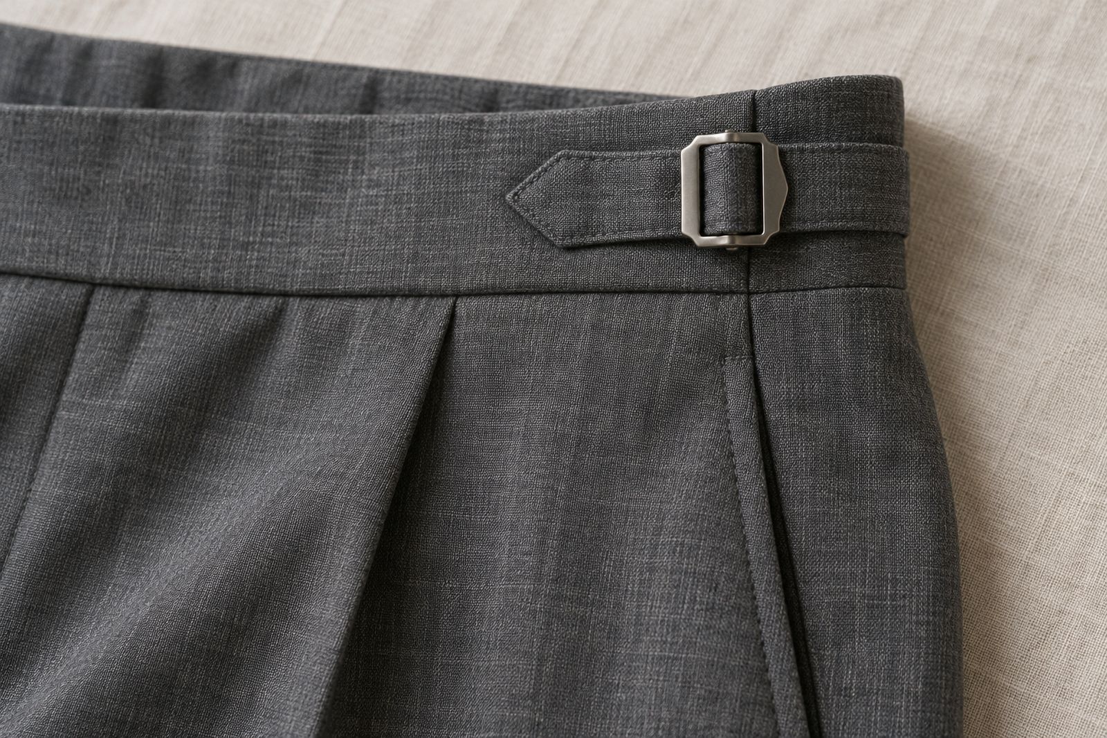 A side adjuster with a metal buckle on the waistband of gray suit trousers with no belt loops.