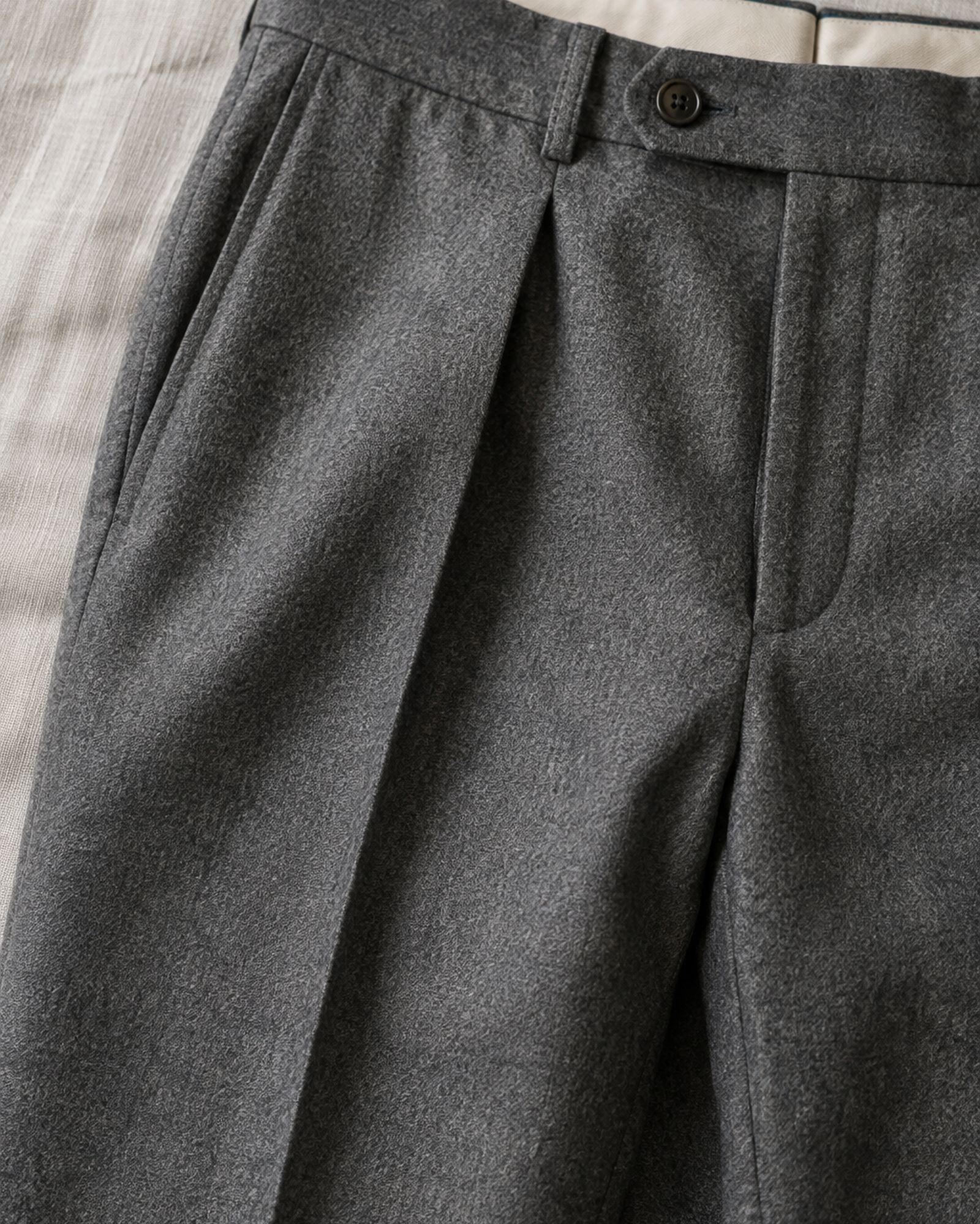 Detail of a forward pleat at the waistband of gray flannel trousers with the crease running down from it.